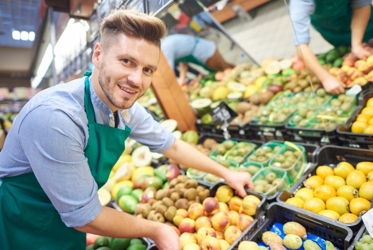 Grocery store employee arranges and restocks the produce section, he is putting a box of nectarines down. He is facing the camera and showcasing how inventory management can help the store.