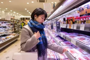 A grocery shopper in a winter coat and scarf shops for meat in a refrigerated aisle, representing the increased seasonal demand grocers face in cold weather.