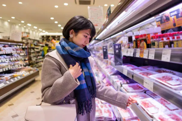 A grocery shopper in a winter coat and scarf shops for meat in a refrigerated aisle, representing the increased seasonal demand grocers face in cold weather.
