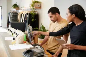 Two retail workers stand at the checkout counter. One points at the computer screen while the other observes, holding a tablet. Clothing and products are displayed in the background.