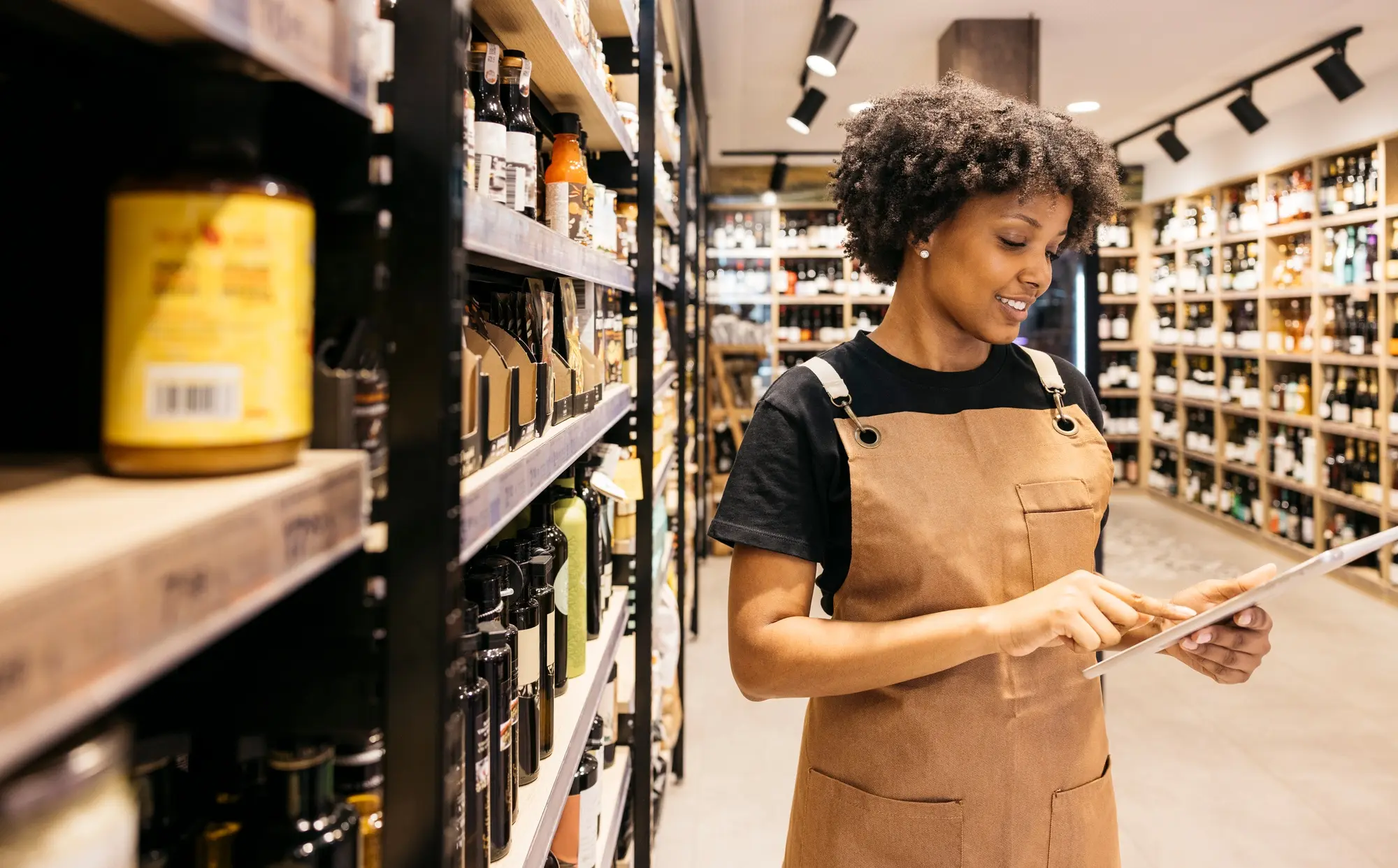 Retail employee in a well-lit store aisle using a tablet to check and manage inventory on neatly stocked shelves.